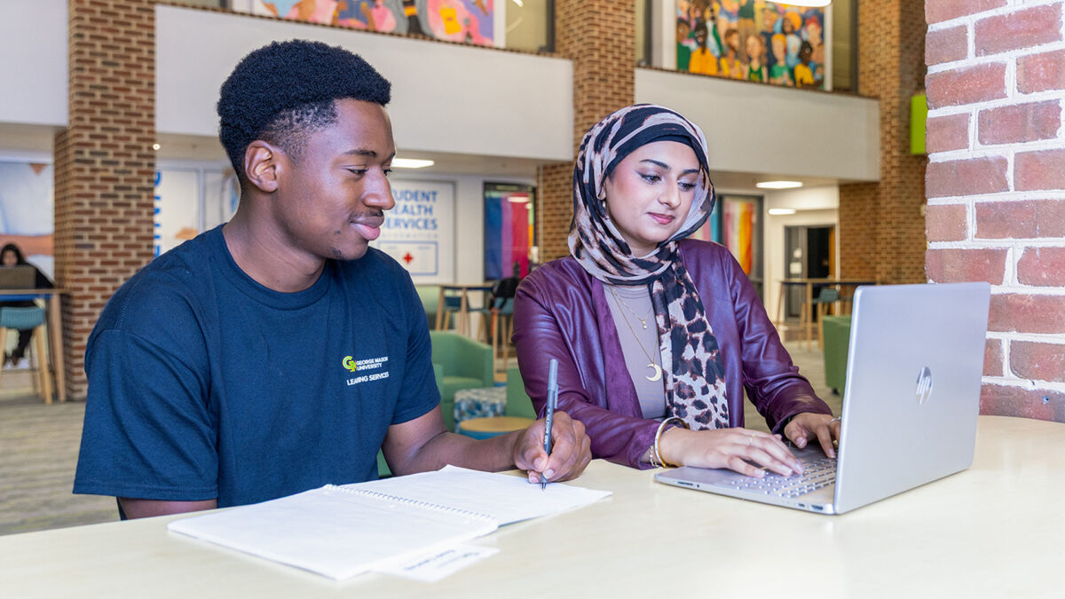 A student with a notebook looks over at their tutor's laptop while smiling.