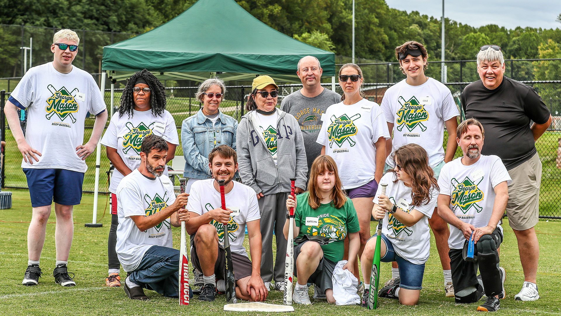 A group of students and staff posing as a team with bats on a field. They wear matching George Mason Beep Baseball shirts.